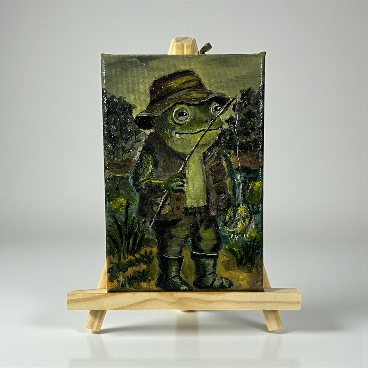 Alkuperäinen öljymaalaus kalastavasta sammakosta Froggies-sarjasta - vihreä sammakko onkivavan kanssa lammen reunalla, käsintehty taideteos