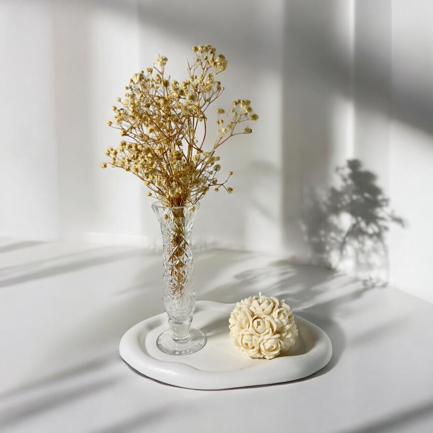 Käsintehty kukkakynttilä ja kuivatut kukat lasimaljakossa, pehmeä valo ja varjot luovat rauhallisen tunnelman vaaleassa tilassa.